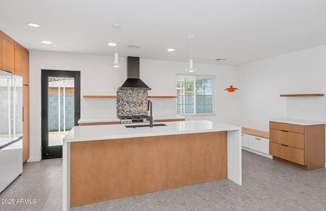 kitchen with pendant lighting, an island with sink, wall chimney range hood, backsplash, and sink