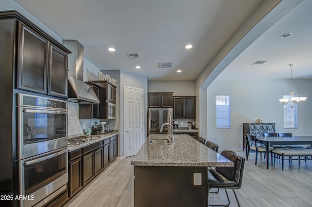 kitchen with wall chimney range hood, sink, a kitchen island with sink, light stone countertops, and stainless steel appliances