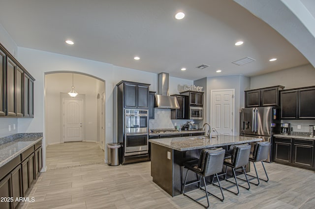 kitchen with an island with sink, appliances with stainless steel finishes, light stone countertops, wall chimney exhaust hood, and sink