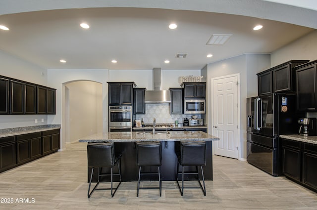 kitchen featuring appliances with stainless steel finishes, backsplash, a kitchen island with sink, wall chimney exhaust hood, and light stone counters