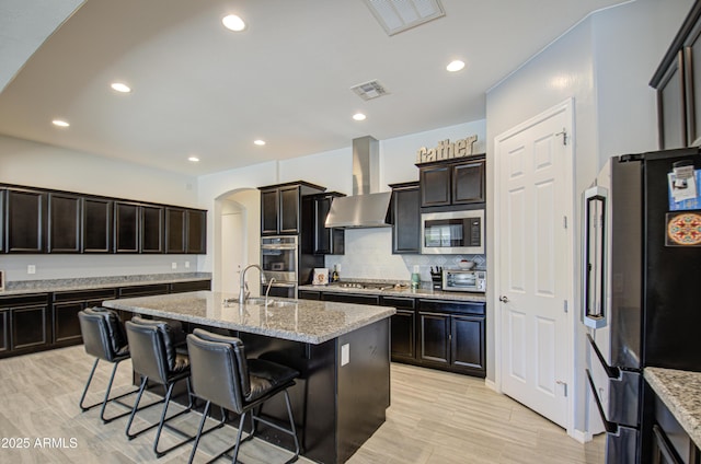 kitchen with wall chimney range hood, a kitchen island with sink, stainless steel appliances, dark brown cabinets, and light stone counters