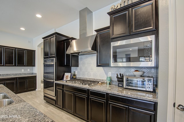 kitchen with backsplash, range hood, light wood-type flooring, light stone countertops, and stainless steel appliances