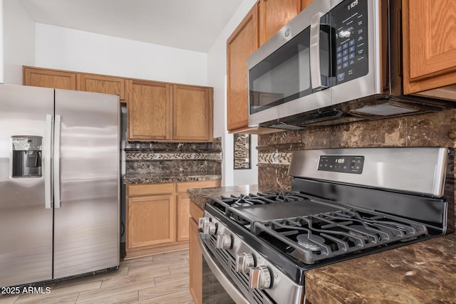 kitchen with dark stone counters, brown cabinets, light wood finished floors, appliances with stainless steel finishes, and backsplash