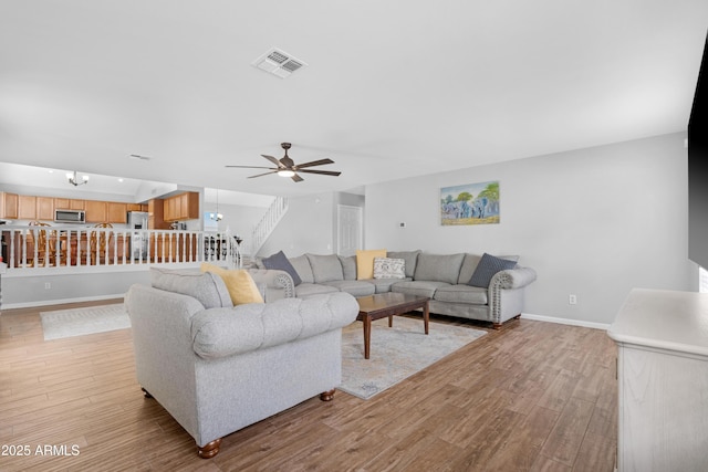 living room featuring visible vents, stairway, light wood-style flooring, baseboards, and ceiling fan with notable chandelier