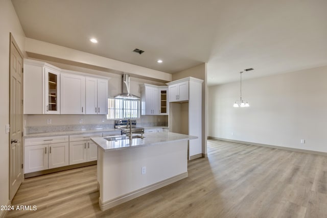 kitchen featuring wall chimney range hood, white cabinets, an inviting chandelier, a sink, and an island with sink