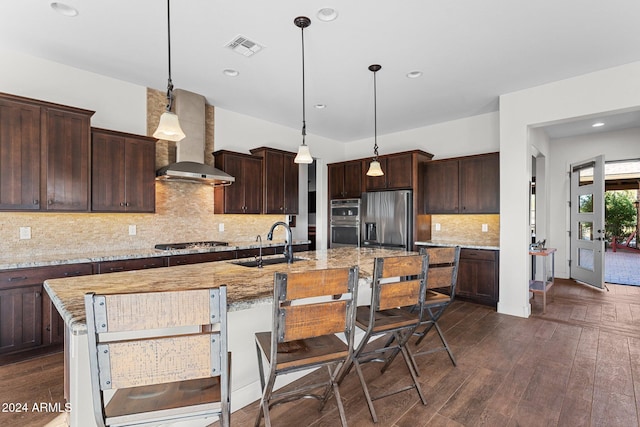 kitchen with dark hardwood / wood-style flooring, light stone counters, wall chimney exhaust hood, stainless steel appliances, and a center island with sink