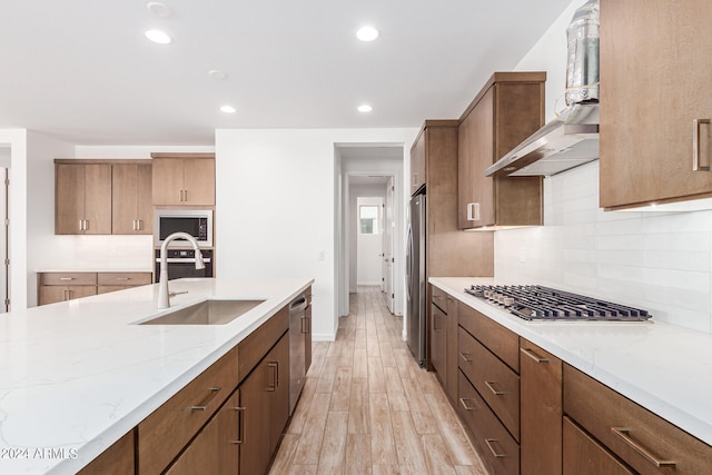 kitchen with light wood-type flooring, light stone counters, stainless steel appliances, sink, and wall chimney exhaust hood