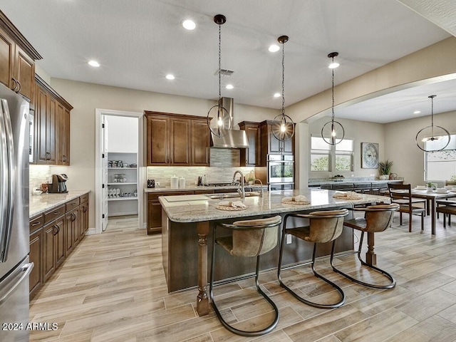 kitchen featuring sink, stainless steel appliances, light stone counters, an island with sink, and decorative light fixtures