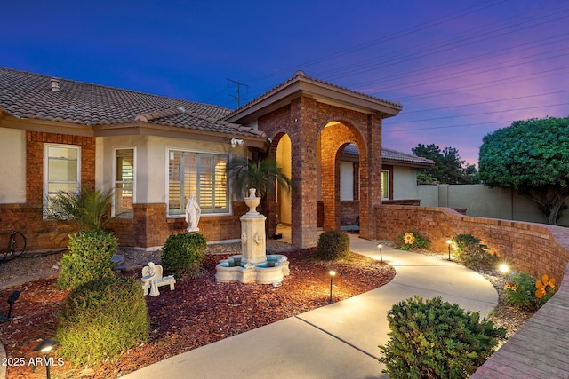 entrance to property featuring brick siding, a tile roof, fence, and stucco siding