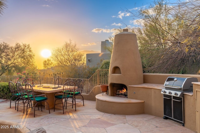 patio terrace at dusk with outdoor dining space, exterior kitchen, a lit fireplace, and fence