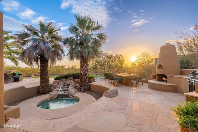 patio terrace at dusk featuring an in ground hot tub, a warm lit fireplace, outdoor dining space, and fence