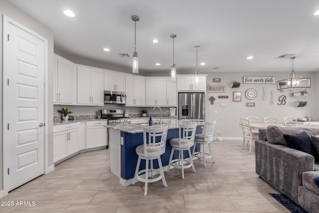 kitchen featuring appliances with stainless steel finishes, a kitchen island with sink, hanging light fixtures, white cabinetry, and light stone countertops