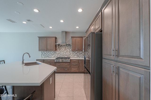 kitchen with sink, black refrigerator, stainless steel gas stovetop, a kitchen island with sink, and wall chimney range hood