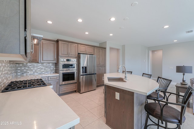 kitchen with appliances with stainless steel finishes, sink, decorative backsplash, light tile patterned floors, and a center island with sink