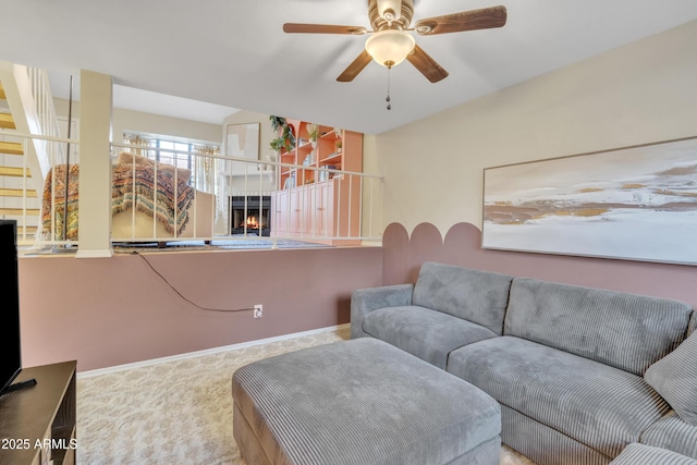 carpeted living area featuring a warm lit fireplace, baseboards, and a ceiling fan