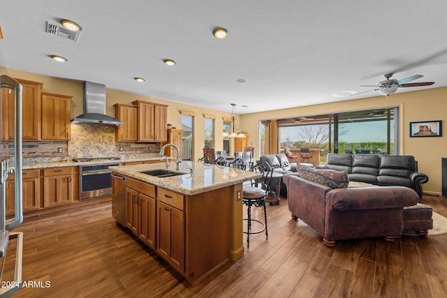 kitchen with sink, appliances with stainless steel finishes, a kitchen island with sink, decorative light fixtures, and wall chimney exhaust hood