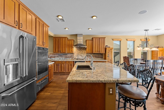 kitchen with sink, a center island with sink, appliances with stainless steel finishes, pendant lighting, and wall chimney range hood