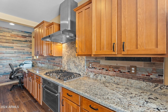 kitchen with appliances with stainless steel finishes, backsplash, dark hardwood / wood-style flooring, light stone countertops, and wall chimney range hood