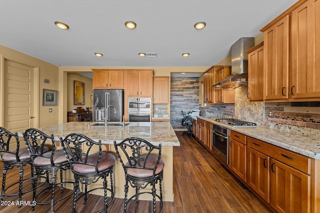 kitchen with appliances with stainless steel finishes, sink, a kitchen island with sink, light stone countertops, and wall chimney exhaust hood