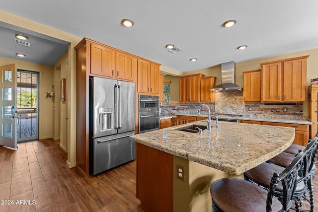 kitchen with wall chimney exhaust hood, sink, a center island with sink, appliances with stainless steel finishes, and light stone countertops