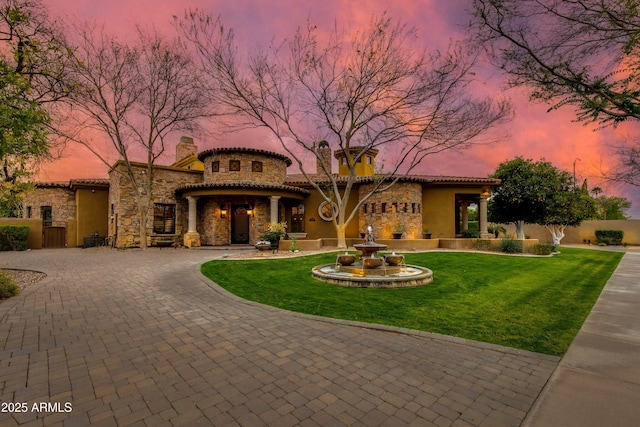 mediterranean / spanish house with curved driveway, a tiled roof, a front yard, stucco siding, and stone siding