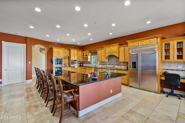 kitchen featuring built in appliances, dark stone countertops, a spacious island, decorative backsplash, and a breakfast bar