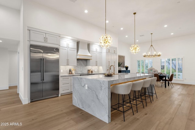 kitchen featuring built in refrigerator, hanging light fixtures, white cabinets, custom exhaust hood, and a large island with sink