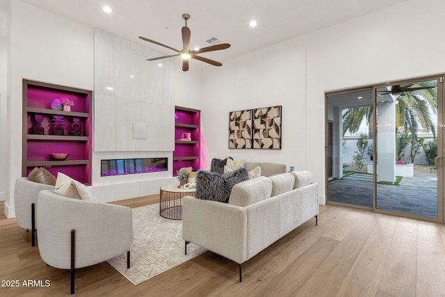 living room with light wood-type flooring, a large fireplace, built in features, a towering ceiling, and ceiling fan