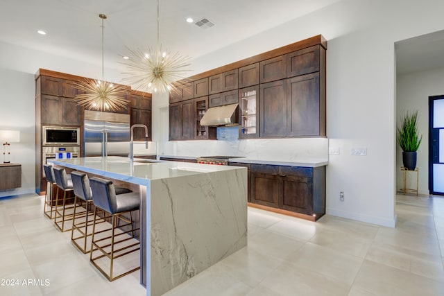 kitchen with ventilation hood, an inviting chandelier, decorative light fixtures, an island with sink, and built in appliances