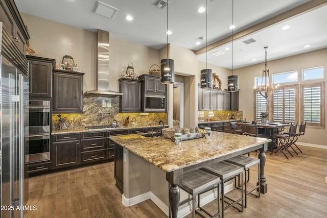 kitchen with an island with sink, light wood finished floors, a breakfast bar area, wall chimney range hood, and visible vents