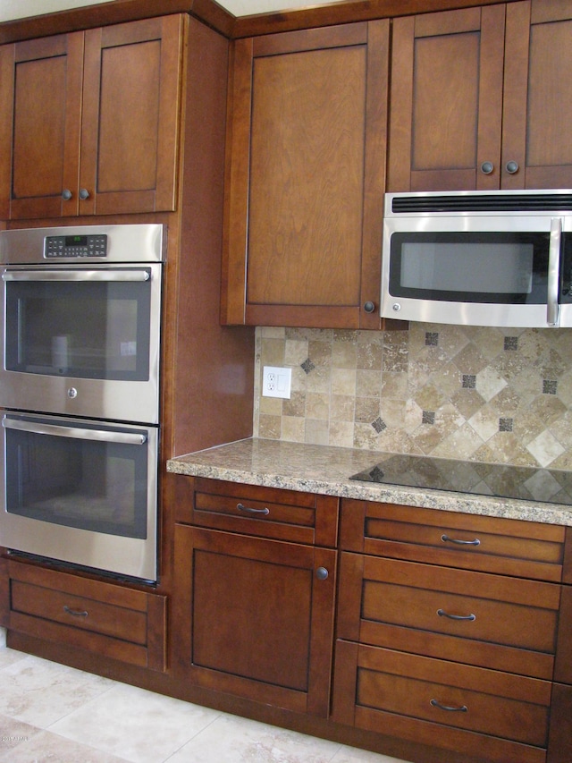 kitchen with light tile patterned floors, brown cabinetry, light stone counters, stainless steel appliances, and backsplash