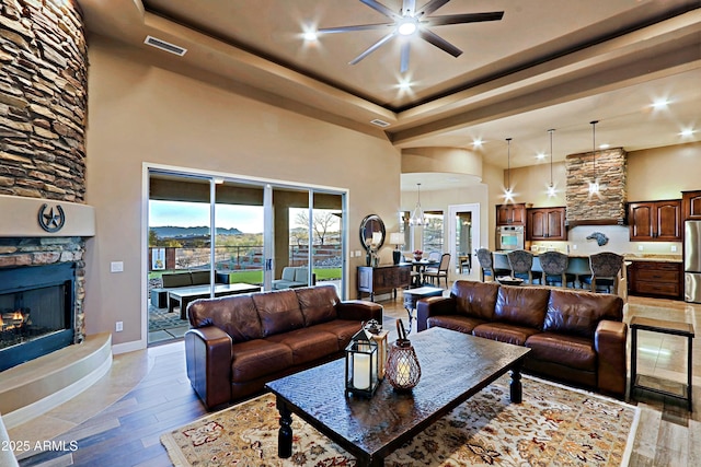 living area with a high ceiling, a glass covered fireplace, a raised ceiling, ceiling fan with notable chandelier, and light wood-type flooring