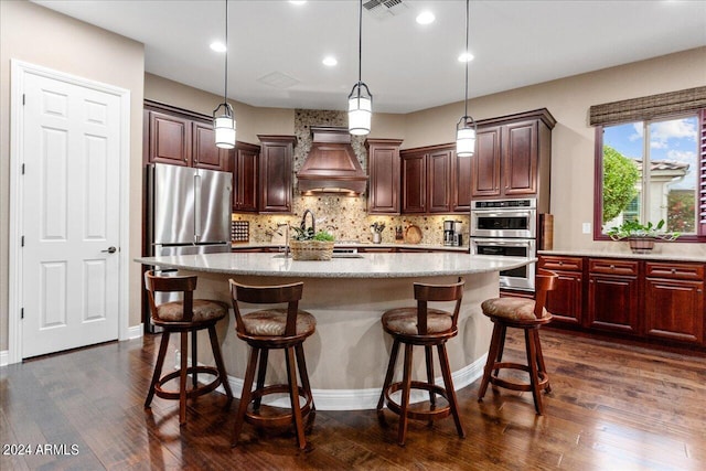 kitchen featuring premium range hood, appliances with stainless steel finishes, a kitchen bar, hanging light fixtures, and a kitchen island with sink