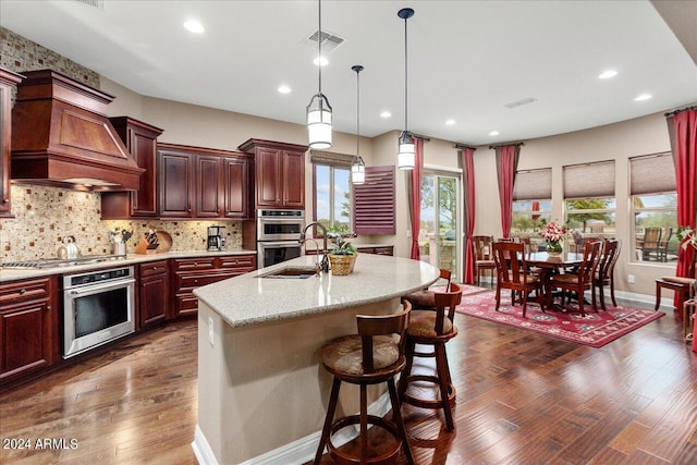 kitchen with premium range hood, decorative backsplash, a kitchen island with sink, and hanging light fixtures