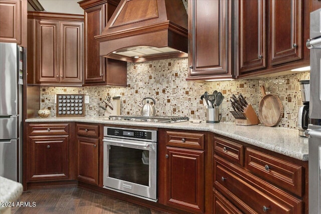 kitchen featuring premium range hood, dark wood-type flooring, light stone counters, appliances with stainless steel finishes, and backsplash