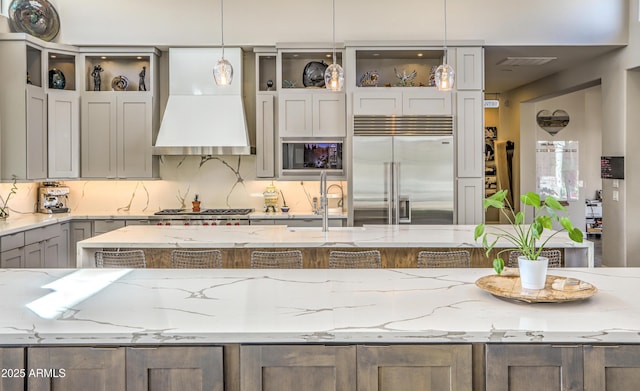 kitchen with light stone counters, pendant lighting, custom range hood, appliances with stainless steel finishes, and a sink