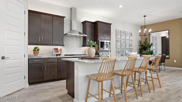 kitchen with appliances with stainless steel finishes, wall chimney range hood, dark brown cabinetry, light hardwood / wood-style floors, and a center island with sink