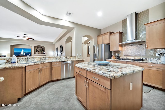 kitchen with ceiling fan, wall chimney exhaust hood, decorative backsplash, a kitchen island, and appliances with stainless steel finishes