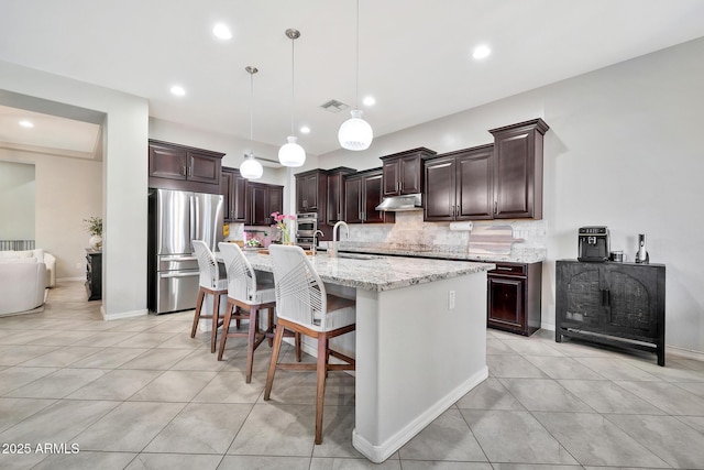 kitchen with light stone counters, dark brown cabinets, a center island with sink, stainless steel refrigerator, and pendant lighting