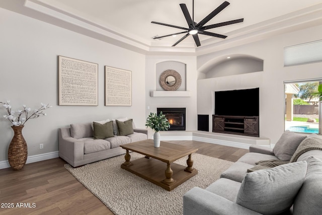 living area featuring ceiling fan, wood finished floors, baseboards, a tray ceiling, and a glass covered fireplace