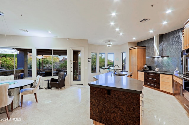 kitchen with tasteful backsplash, stainless steel appliances, a kitchen island with sink, sink, and wall chimney range hood