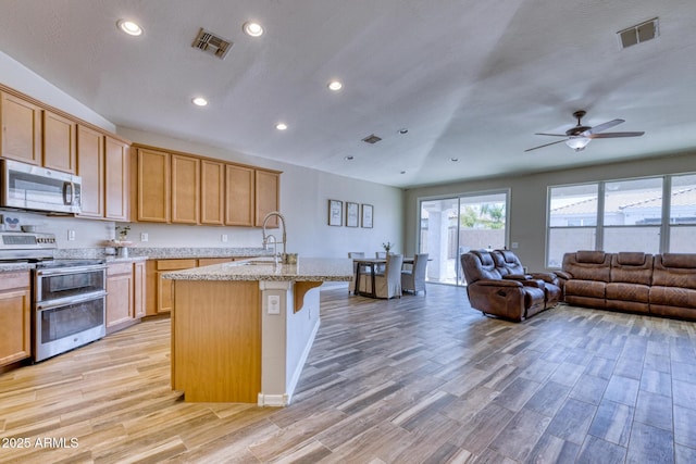 kitchen with sink, appliances with stainless steel finishes, light stone countertops, an island with sink, and light wood-type flooring