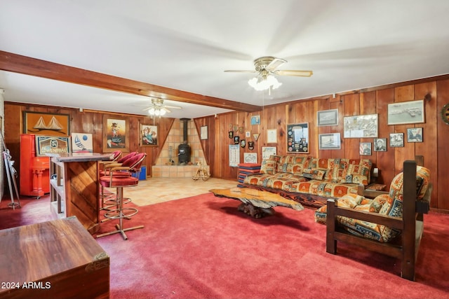 carpeted living room featuring wood walls, a wood stove, ceiling fan, bar, and beamed ceiling