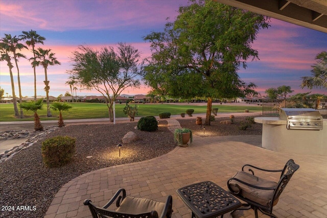 patio terrace at dusk with area for grilling, a yard, and exterior kitchen