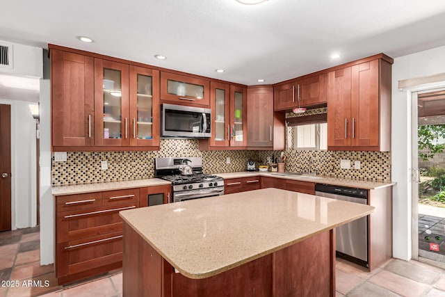 kitchen featuring light stone counters, a sink, stainless steel appliances, decorative backsplash, and glass insert cabinets