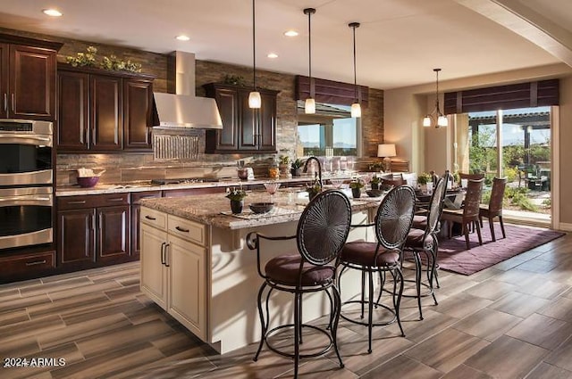 kitchen with hanging light fixtures, stainless steel appliances, wall chimney range hood, a kitchen breakfast bar, and a kitchen island with sink