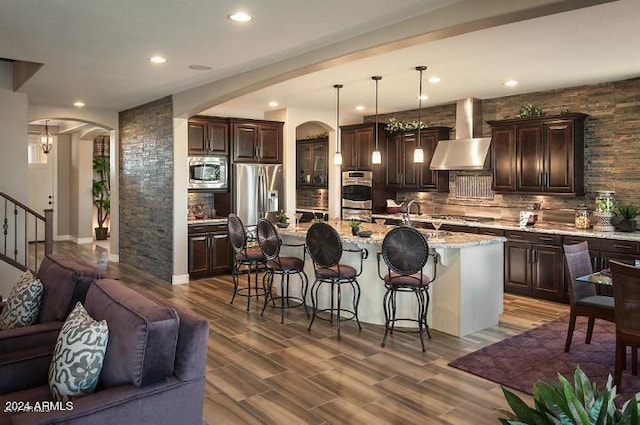 kitchen with arched walkways, open floor plan, stainless steel appliances, wall chimney range hood, and a kitchen bar