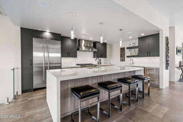 kitchen with a kitchen island with sink, tasteful backsplash, built in refrigerator, decorative light fixtures, and wall chimney exhaust hood