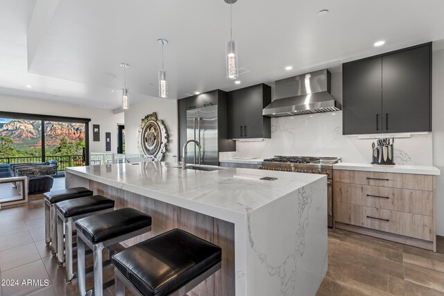 kitchen featuring hanging light fixtures, light stone counters, a large island with sink, wall chimney exhaust hood, and a kitchen breakfast bar