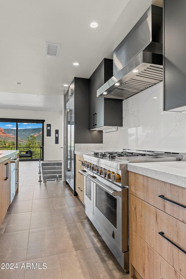 kitchen featuring range with two ovens, light brown cabinetry, light tile patterned floors, light stone counters, and wall chimney range hood